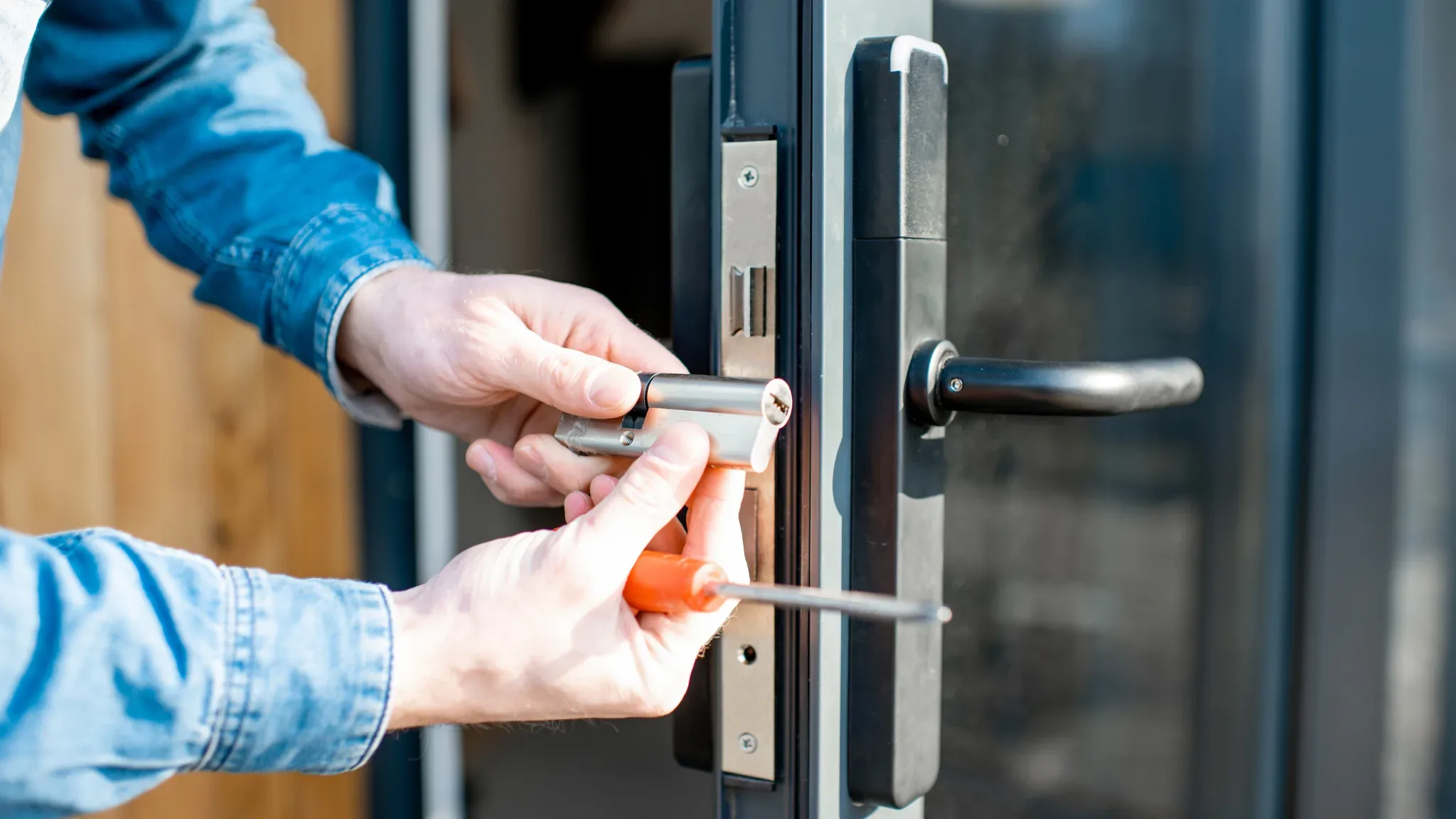 An extreme close-up shows two hands installing a euro-cylinder lock into the edge of a dark-framed glass door: one hand holds