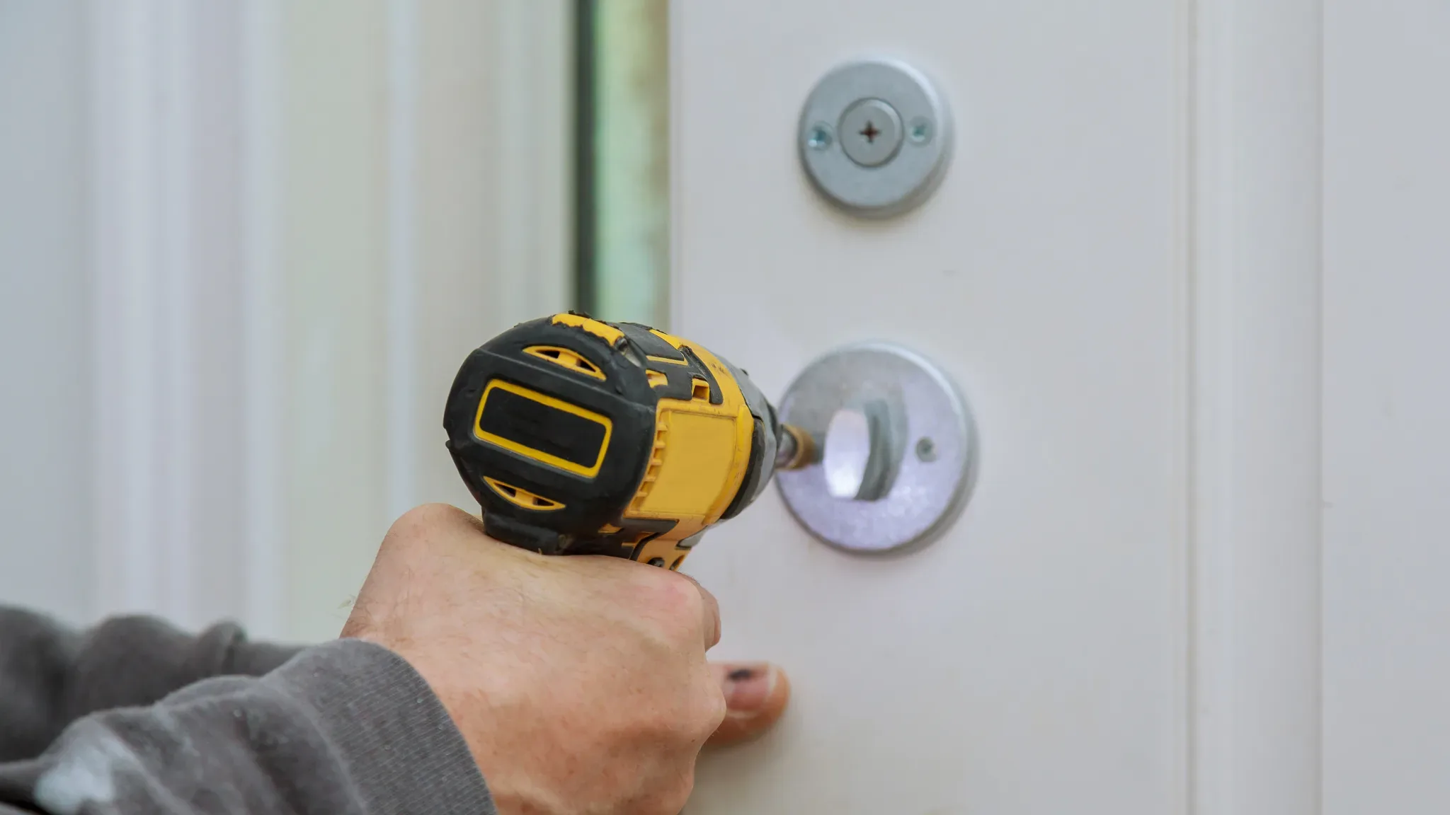 A hand grips a black and yellow impact driver, its bit inserted into a round silver deadbolt cylinder mounted on a white inte